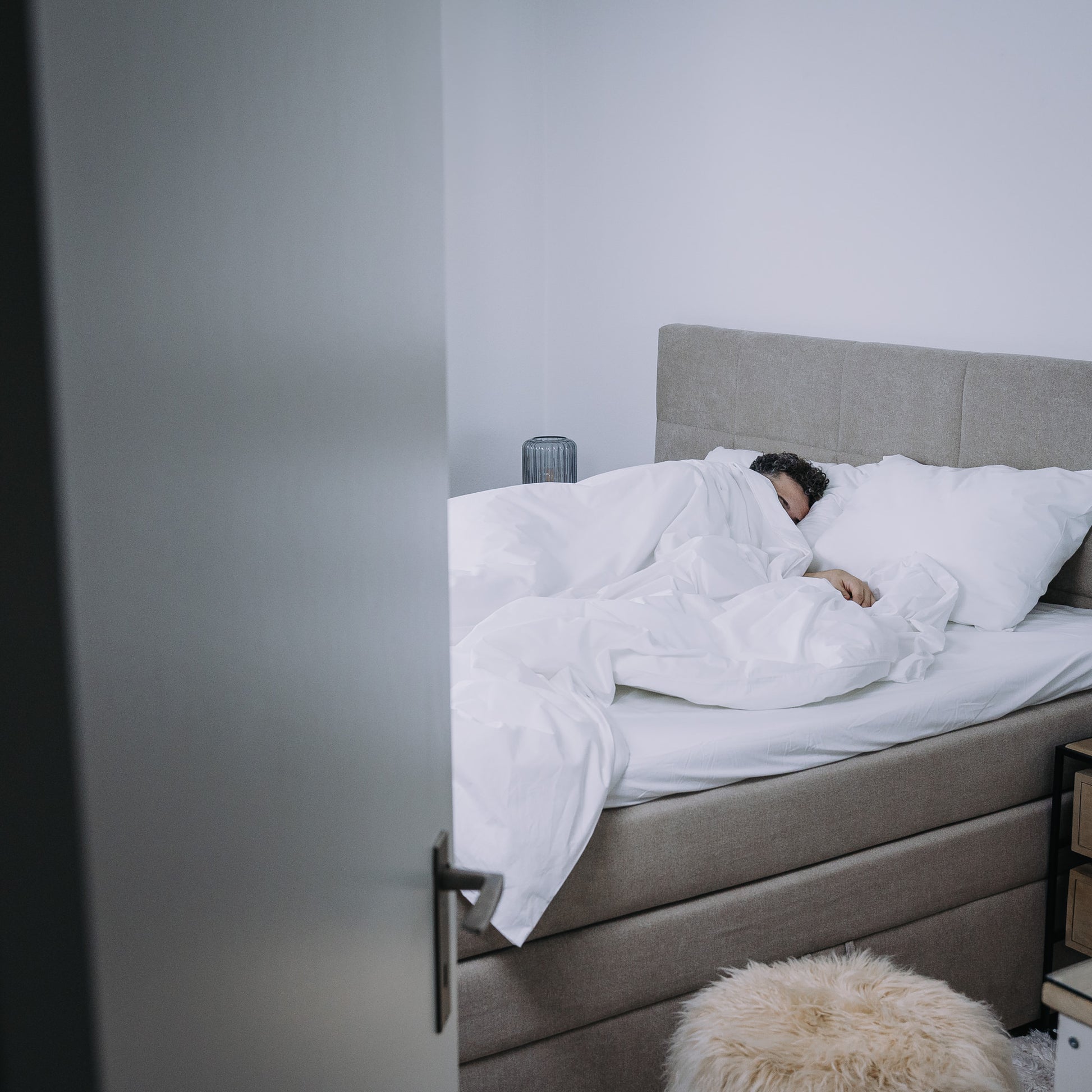 Person lying in bed under white duvet with gray headboard and pillows.